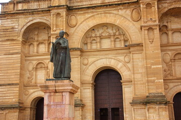 Statue of a robed figure on a stone pedestal in front of an ornate sandstone building with arched doorways, intricate carvings, and dark wooden doors;