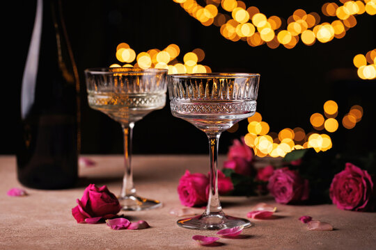 Two empty crystal coupe glasses with pink roses and warm fairy lights in the background, evoking a celebratory mood.