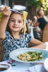 A little girl happily digging into a bowl of pasta with green pesto sauce and arugula, surrounded...