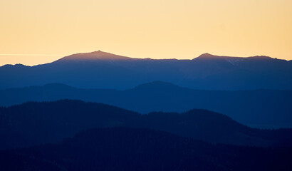 Silhouette of distant mountains at sunset, with golden sky transitioning into deep blue shadows. Ridges and peaks create stunning contrast against warm hues of horizon.