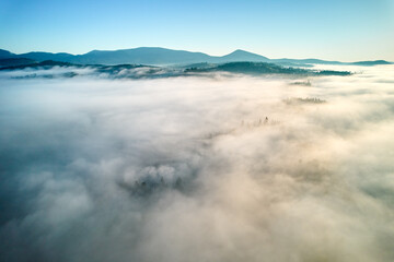 Aerial view of landscape enveloped in thick, rolling clouds. Verdant hills and scattered trees barely peek through, while backdrop of distant mountains completes this serene and dreamlike scene.