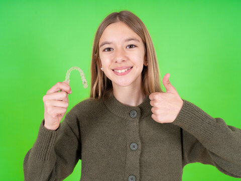 beautiful caucasian girl wearing green sweater holding an invisible braces aligner and rising thumb up, recommending this new treatment. Dental healthcare concept.