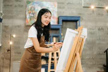 Cheerful female artist painting in a vibrant studio with brushes, a palette, and a blank canvas.