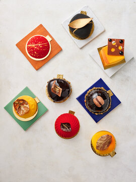 Various elegant desserts are arranged on colorful square plates, showcasing intricate designs and bright colors. The shop setting emphasizes a creative pastry presentation