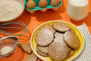 Close up of oatmeal cookies sprinkled on a yellow plate against an orange background next to cooking materials. Cookies made from wheat flour mixed with oat cereal, a traditional pastry