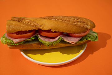 Sandwich with bread, lettuce, ham, and tomato on a yellow plate against an orange background. Sandwich on a colorful background