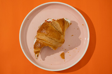 Top view of a half moon croissant filled with cream cheese and strawberry jam, with a bite taken out. On a pink plate against a minimalist orange background. Puff pastry. Bakery item