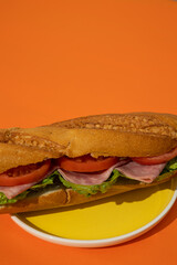 Vertical image of a sandwich with bread, lettuce, ham, and tomato on a yellow plate against an orange background. Sandwich on a colorful background