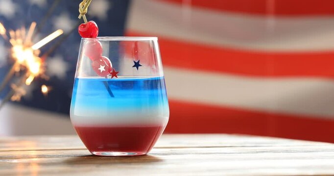 Woman adding maraschino cherries to cocktail at wooden table against American flag, closeup. Space for text