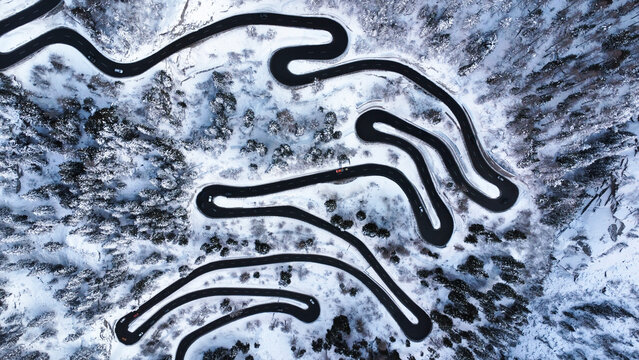 Aerial view of winding road through a snowy landscape with many little cars tackling the treacherous Maloja Pass, Graubuenden Canton, Switzerland.