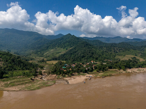 Aerial view of a picturesque rural landscape featuring a serene river, lush greenery, and a tranquil village surrounded by mountains and forests, Pak Tha District, Bokeo, Laos.