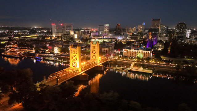 Aerial view of illuminated tower bridge and vibrant cityscape at night with reflections on the river, The Bridge District, West Sacramento, California, United States.