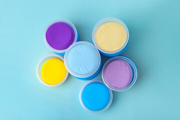 Different colorful modeling clay in plastic jars on light blue background, flat lay