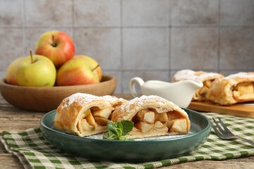 Pieces of tasty apple strudel with powdered sugar and mint on wooden table against grey tiled wall, closeup