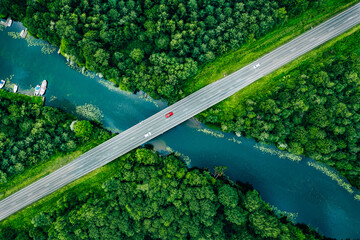 Aerial view of asphalt road with white and red cars over blue river and green summer woods in Finland, drone photography