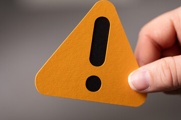Close-up of a hand holding an orange triangle warning sign with an exclamation mark.