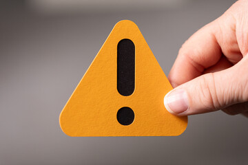 Close-up of a hand holding an orange triangle warning sign with an exclamation mark.