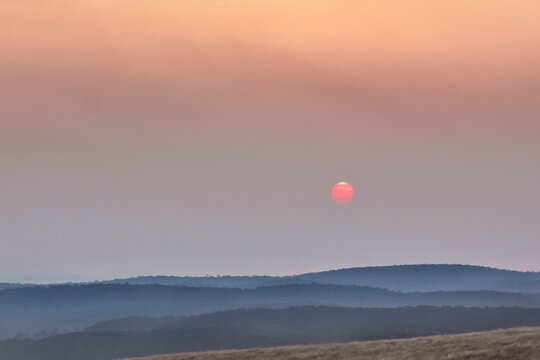 A red Sun sinking into smoke haze over distant mountains