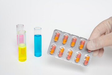 Gloved hand holding a blister pack of orange and yellow capsules with colorful test tubes on isolated white background. Medical research with pills and vials. Science, medicine and laboratory concept