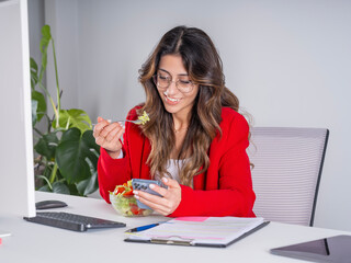 Businesswoman eating salad,  young caucasian happy smiling businesswoman eating salad during lunch break sitting desk in office looking smartphone screen using mobile phone. Copy space.
