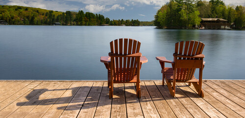 Two Adirondack chairs rest on a wooden dock, overlooking a tranquil summer lake in Muskoka. The...