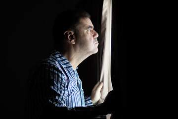 A frowning man in striped pajamas watches the street from behind a window curtain