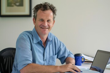 Happy smiling male sitting at desk in coworking office, drinking coffee from takeaway cup and doing work on his laptop, looking at camera