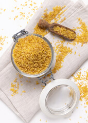 Glass jar of raw dry uncooked yellow bulgur grain with a wooden scoop on napkin top view