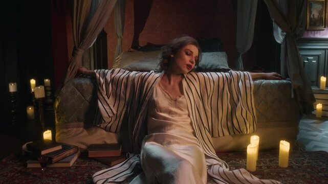 High angle shot of vampire woman in flowing white dress and striped robe leaning back against bed, surrounded by candles, scattered books, patterned rug, and cozy curtains in warmly lit room
