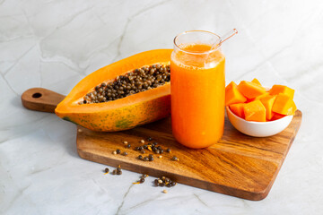 A halved papaya, vibrant smoothie in a glass jar with a straw, fresh cubes in a bowl, and scattered...