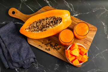 A halved papaya, vibrant smoothie in a glass jar with a straw, fresh cubes in a bowl, and scattered...