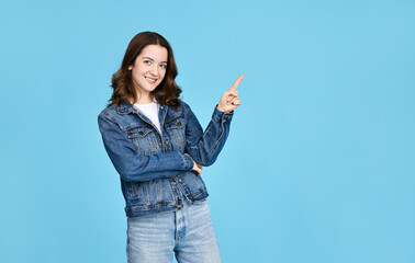 Portrait of cute girl in denim jacket pointing finger to the side presenting advertisement. Isolated on blue background with free space.