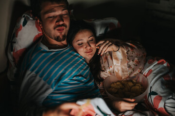 A couple relaxes in bed, sharing a joyful moment with snacks. The warm lighting creates a cozy,...