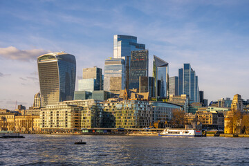 Obraz premium Modern skyscrapers rise against a clear sky in London City, reflecting on the River Thames. Boats glide along the water, showcasing the vibrant urban landscape of England.
