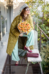 Trendy woman in a chic outfit poses with a bouquet of tulips on a stylish patio. Dressed in a green coat, striped pants, and red heels, she exudes confidence and elegance in an urban nature setting