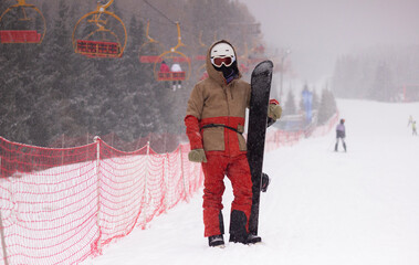 snowboarder standing next to a chairlift holding a snowboard, snowy weather, ski resort, snowboard...