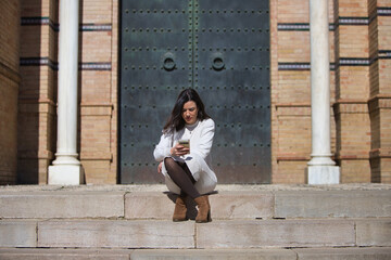 Young and beautiful Latin brunette woman is sitting on a staircase looking at her mobile phone. The...