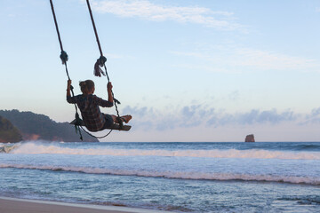 Happy girl have fun swinging high in mid air. Flying on rope swing on sea beach. Travel adventure on paradise tropical island. Family lifestyle, activity on summer vacation.