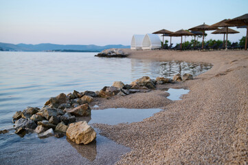 Calm waters reflect the evening sky while rocks line the sandy shore where beach umbrellas stand.