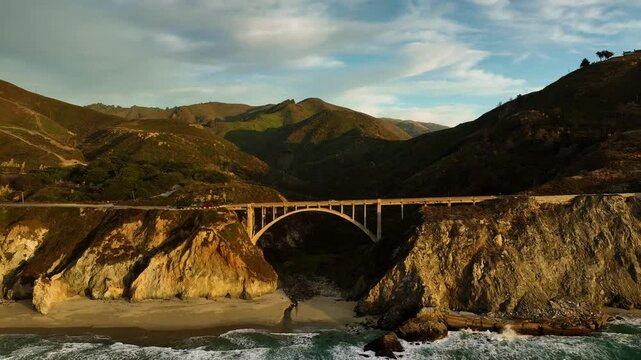 Aerial view of bixby creek bridge at sunset with waves crashing against rocky cliffs, big sur, california, united states.