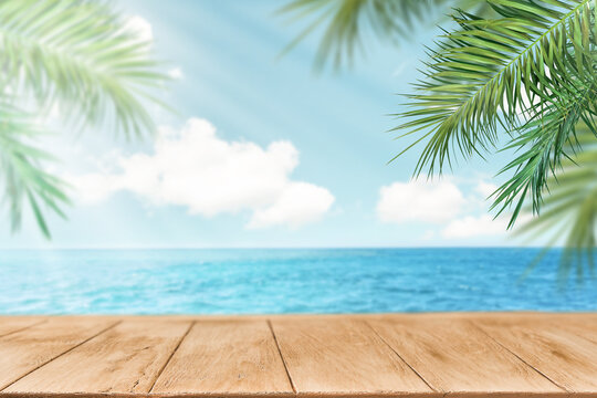 Summer sea and seascape with palm branches, sun and clouds with empty wooden table space for text. Defocus background. Blurred travel and vacation backdrop. Abstract summer background design element.
