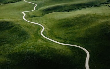 Fototapeta premium Aerial view of a winding dirt road traversing lush green hills. The road curves gently through the landscape, creating a visual sense of journey and exploration