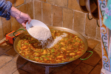 Man's hand adding rice with a bowl to a paella pan, slowly cooking the sauce with vegetables and meat to prepare the authentic Valencian paella in a rustic kitchen with a butane gas grill.