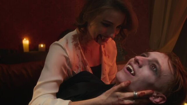 Pale vampire woman with fangs and blood smeared on her face and open smile, leaning over her newborn vampire lover amidst flickering candlelight