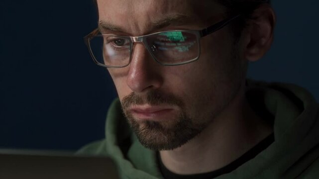 A male programmer is working late, analyzing data on his monitor. His glasses reflect the programming script as he focuses on cybersecurity and machine learning.
