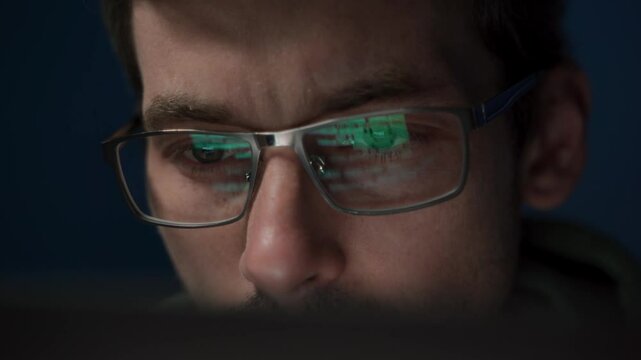 Close-up of a coder in glasses, intently working on a script. The reflection of programming code is visible on his eyeglasses as he develops an application.