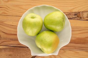 Several sweet green apples in a ceramic plate on a wooden table, close-up, top view.