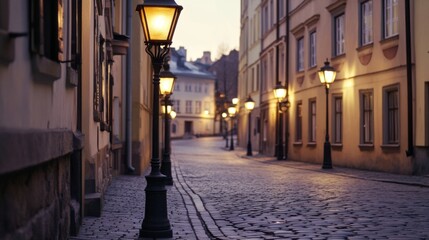 Naklejka premium Vintage-style street lamps lining a cobblestone street, softly glowing in the early evening light, with old buildings in the background.