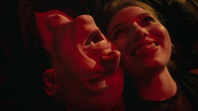 Close-up high angle shot of smiling vampire man and woman lying face-to-face in warm, red-tinged dim light