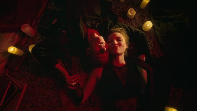 Top view shot of two smiling vampire lovers, hand-in-hand and wearing dark clothes, lying on patterned rug amidst the warm, reddish glow of numerous lit candles